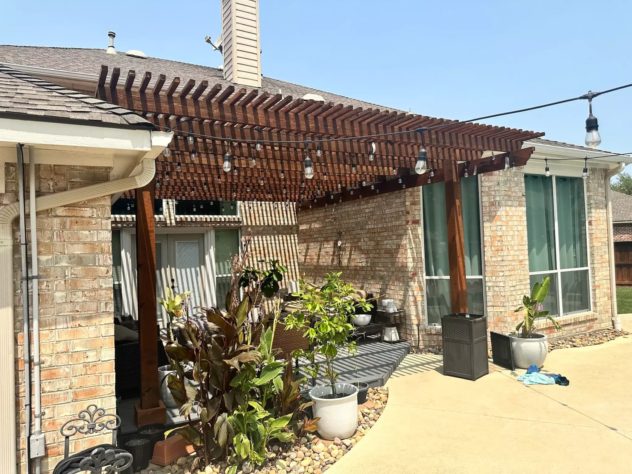 Dark brown stained wooden pergola in a home's backyard alcove, with lights strung across it and greenery beneath it