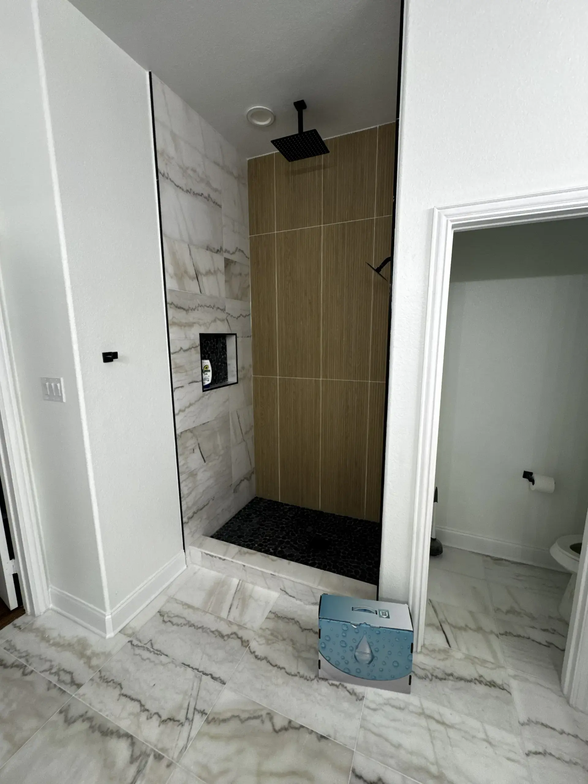 Modern angular shower with seamless marble tiles, wood accents, a dark floor and shelf, and a black stainless steel rain shower head in the ceiling