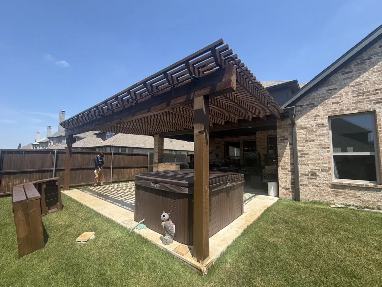 Dark brown stained wooden pergola in a home's backyard alcove, with lights strung across it and greenery beneath it